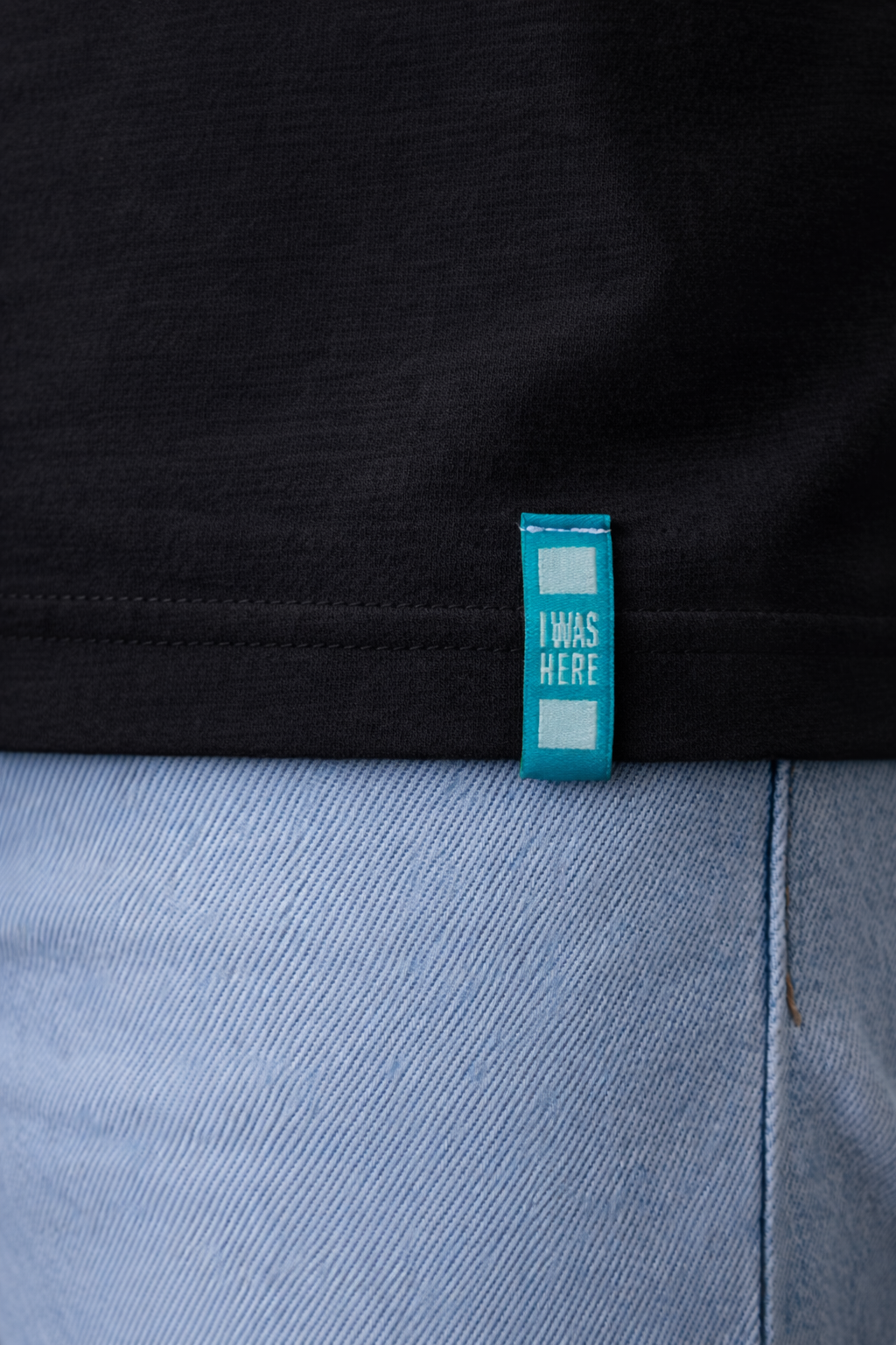 Detailaufnahme eines schwarzen Premium T-Shirts mit türkisfarbenem I WAS HERE Hem-Tag am Saum