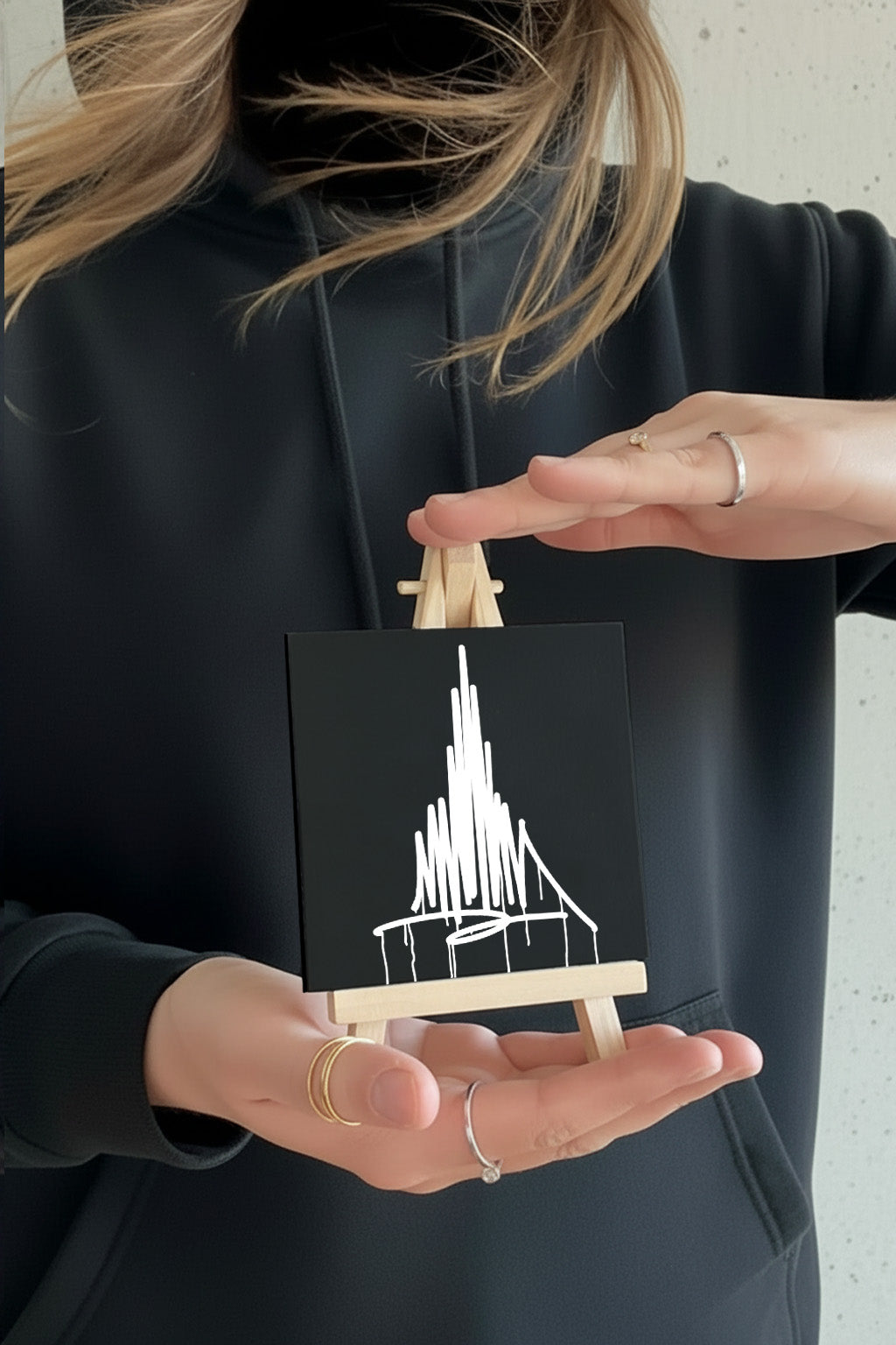 Kleine schwarze Designspiegel-Tafel mit weißem Münster-Urban-Art-Motiv auf Mini-Staffelei, von einer Person im Hoodie in den Händen gehalten.