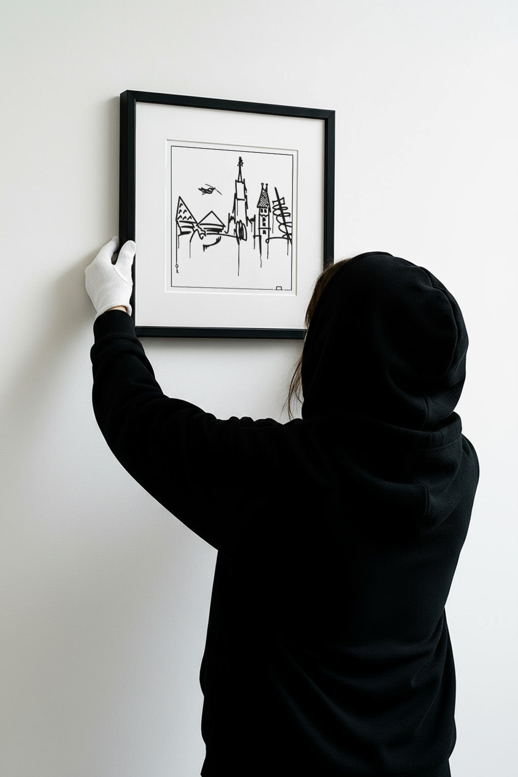 Seitliche Aufnahme einer Person im Hoodie, die einen gerahmten Ulm Skyline Kunstdruck an die Wand hängt – moderner urbaner Wohnstil.
